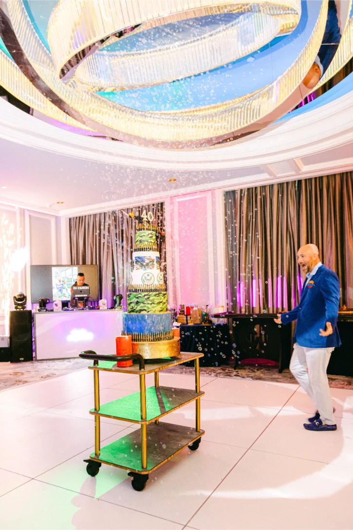 Formally-dressed man with excited expression, walking towards birthday cake. DJ CERON plays music behind him.